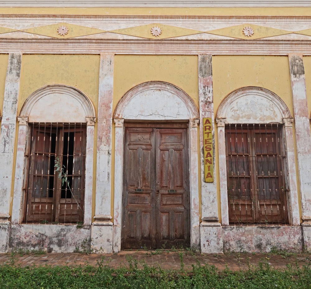 The facade of an old plastered building, its arched doorway flanked by shuttered windows and a hand-painted ARTESAN&Iacute;A sign