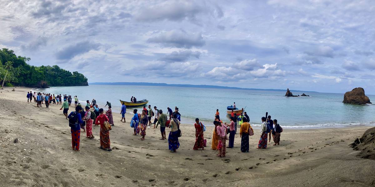 A large group of people walking along a curving beach towards two moored boats, with forested headland behind and rock stacks offshore.