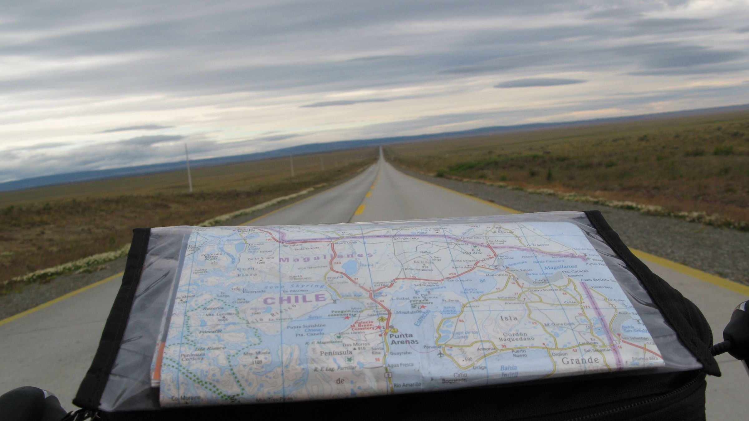 A cyclist's view over a handlebar bag holding a folded road map, looking down a long straight road running to the horizon across open steppe under a broken grey sky.