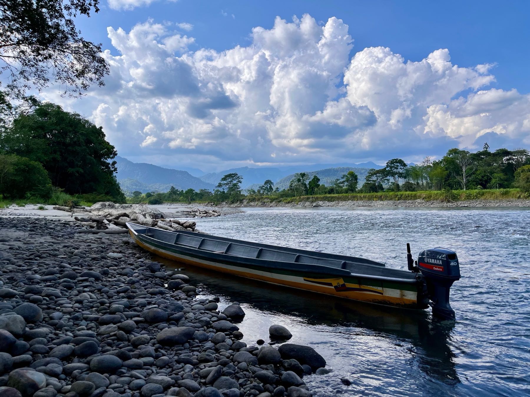 A motorised dugout canoe pulled up on a cobbled river bank, with forested banks and mountains beyond.
