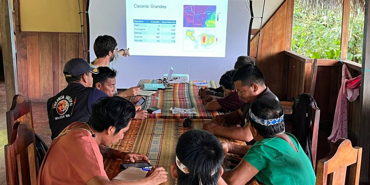 People seated along a long table in an open-sided wooden building, watching a projected slide of hunting counts and habitat maps.