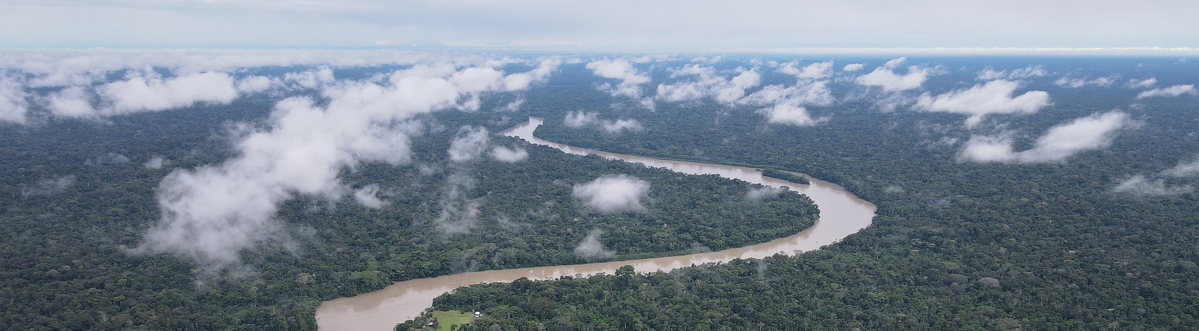 Aerial view of a wide brown river meandering in long bends through unbroken lowland forest, with low cloud sitting on the canopy and a small riverside clearing near the near bank.
