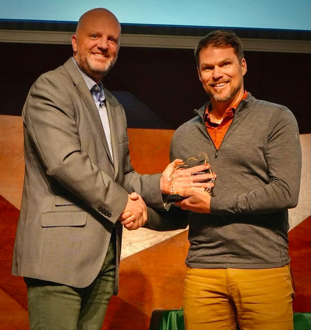 Joel E. Correia and another man shaking hands on a stage, both smiling toward the camera, holding a glass award between them.
