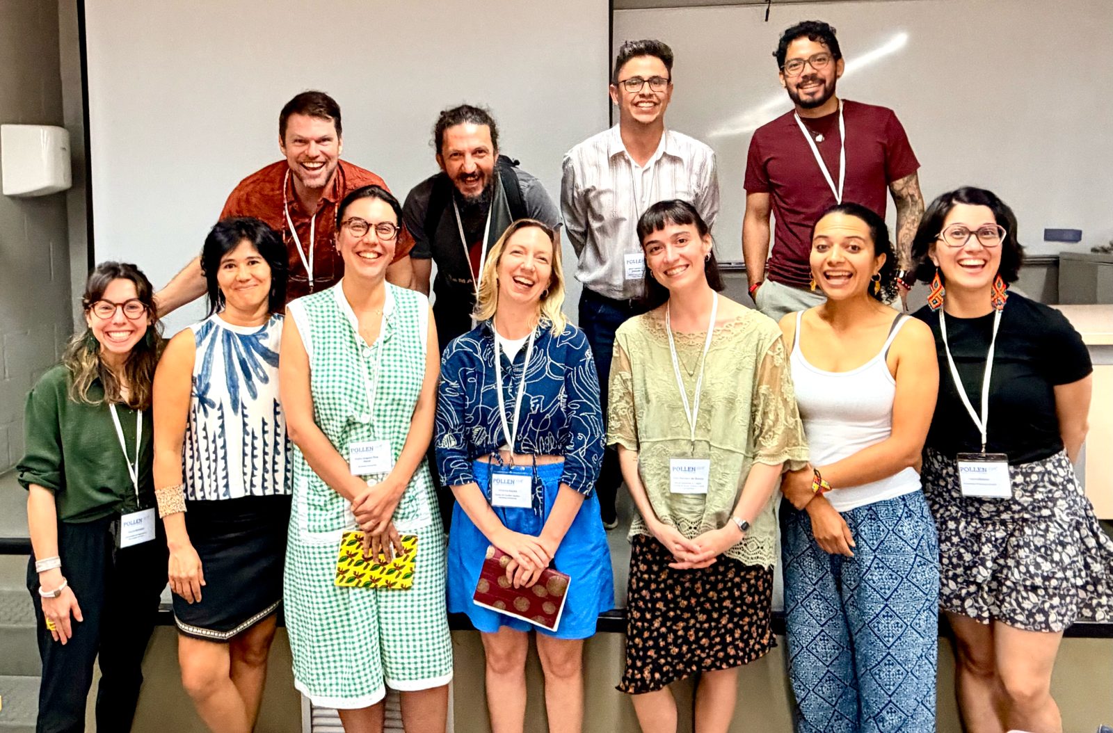 Eleven people standing together in two rows at the front of a lecture room, smiling at the camera, most of them wearing POLLEN conference lanyards.