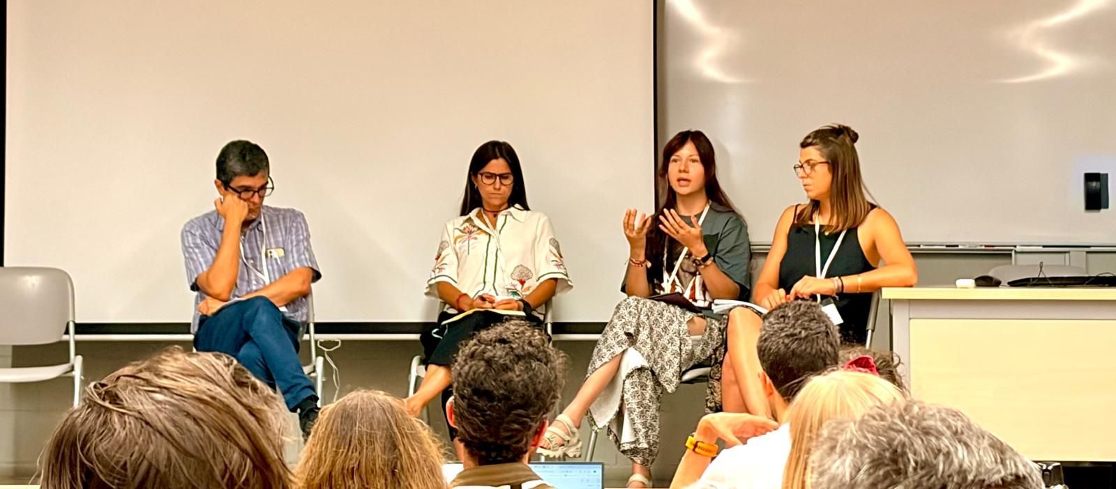 Four panelists seated at the front of a lecture room, one of them speaking with both hands raised, seen over the heads of the audience.