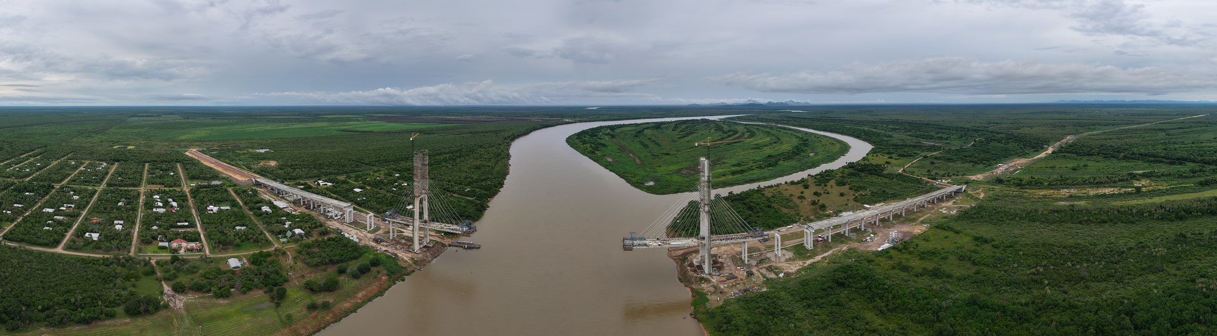 Aerial panorama of a cable-stayed bridge under construction across the Río Paraguay, its two pylons and approach viaducts rising from cleared ground on either bank, with a riverside settlement on the left and forest and pasture running to the horizon.