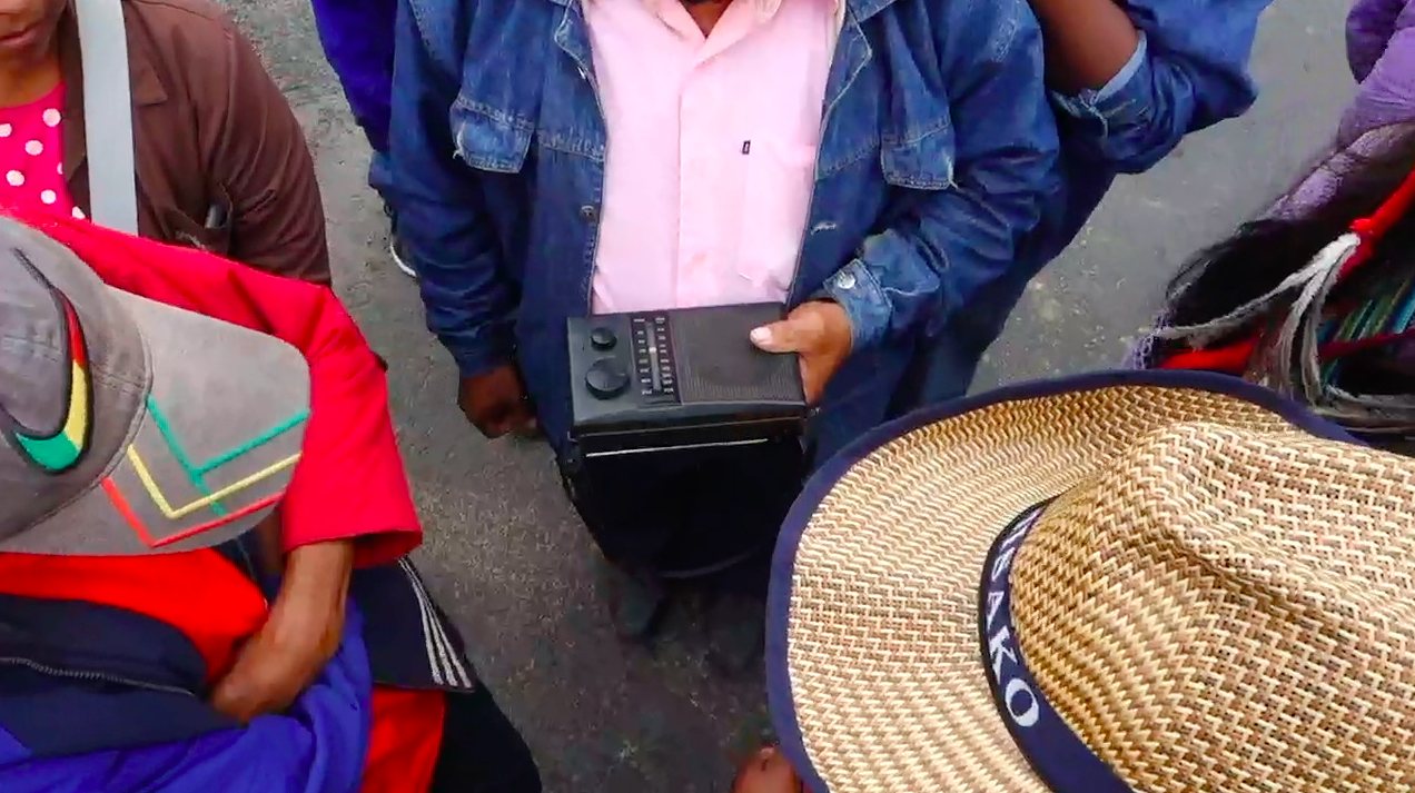 A group of people standing close together outdoors, one of them holding a portable transistor radio at waist height; woven and brimmed hats in the foreground.